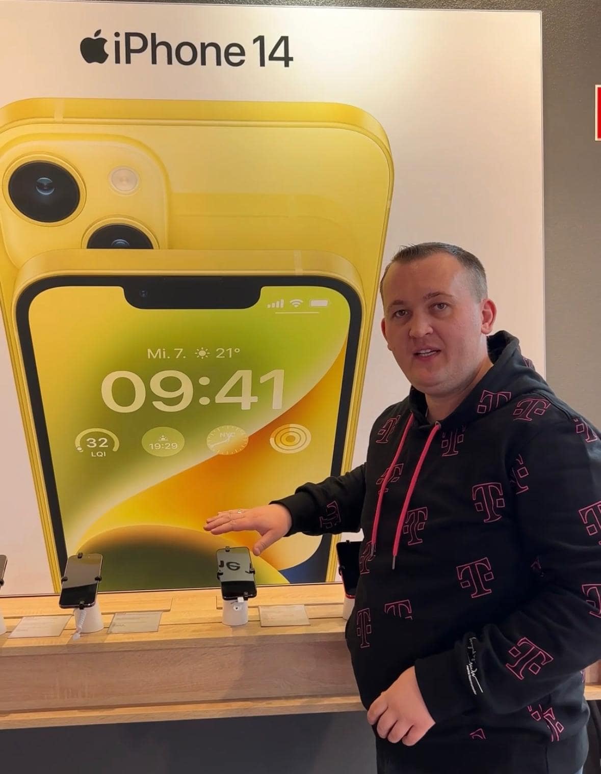 Telekom Shop Mitarbeiter steht vor einem Plakat, auf dem ein iPhone 14 zu sehen ist.