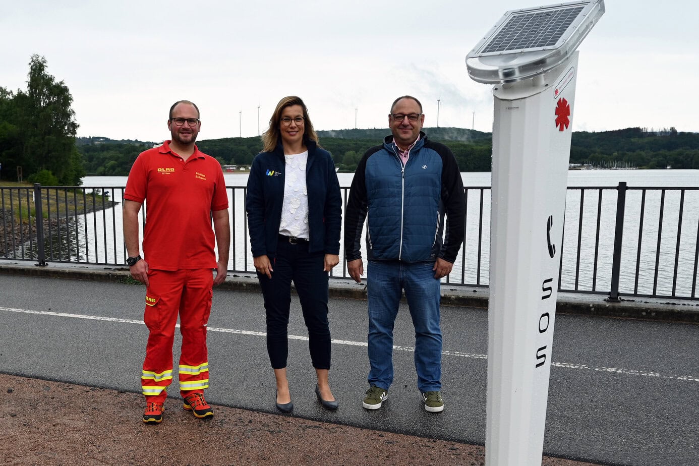 Philipp Scherer (stellvertretender Leiter Einsatz der DLRG und Beauftragter Wasserrettungsdienst Stationär), Daniela Maslan-Mayer (Leiterin Freizeitzentrum Bostalsee), Michael Geisenhainer (Vorsitzender Tauchclub St. Wendel) an der Notrufsäule am Seerundweg an der Staumauer. Fotografin: Inga Schmidt