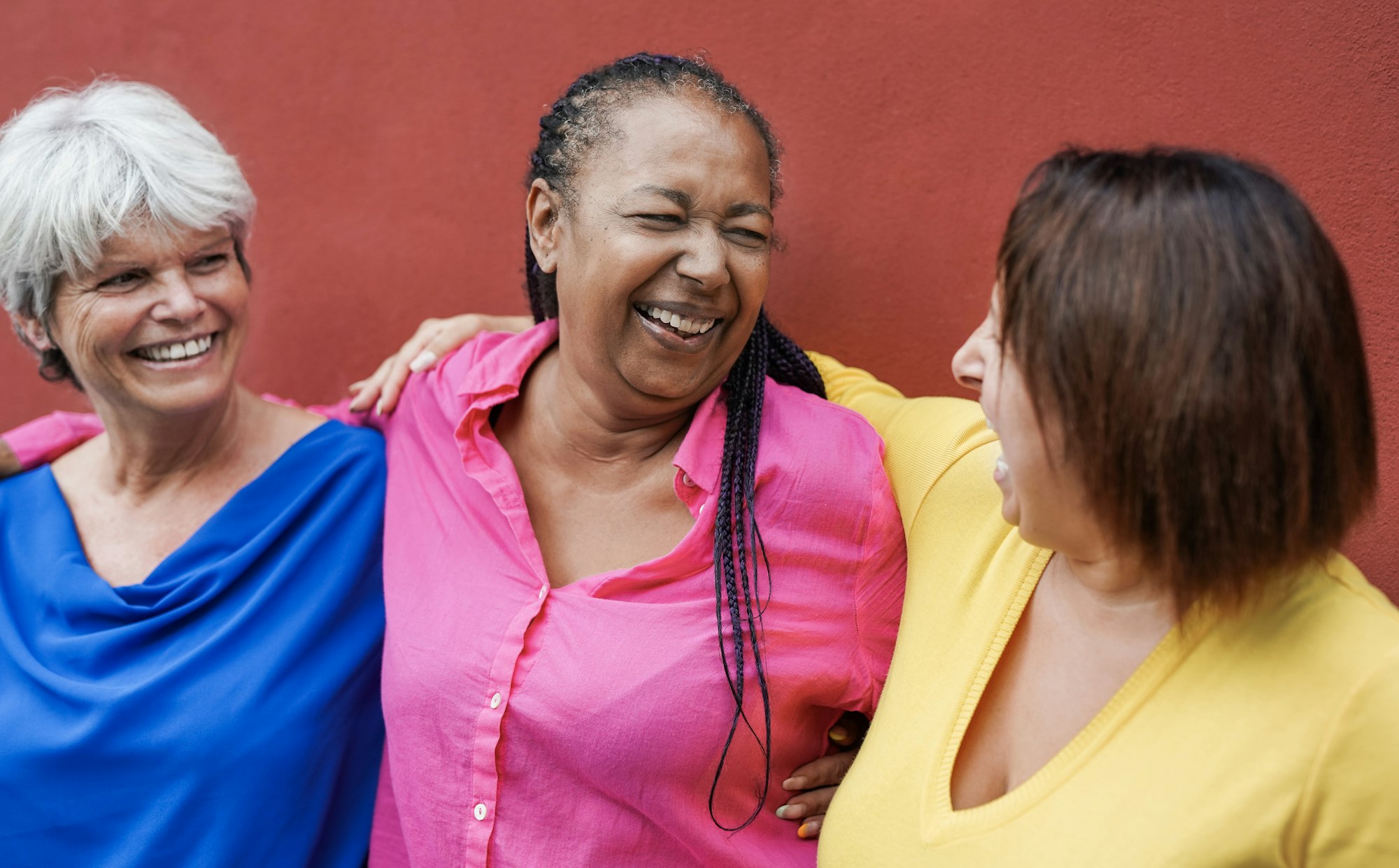 Multiracial mature women hugging each other in the city with red wall in background