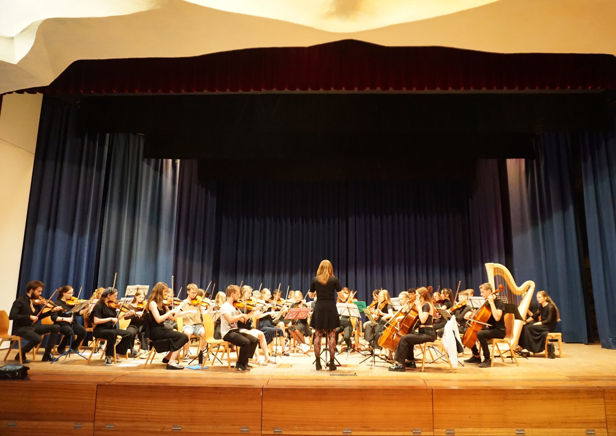 Bühne mit verschiedenen Schülern und einer Dirigentin, welche musizieren.