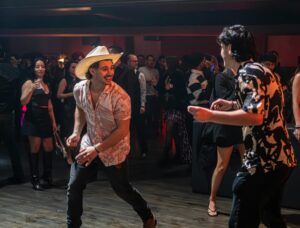 mehrere Tanzendende Menschen mit Cowboyhüten in einem dunklen Saal