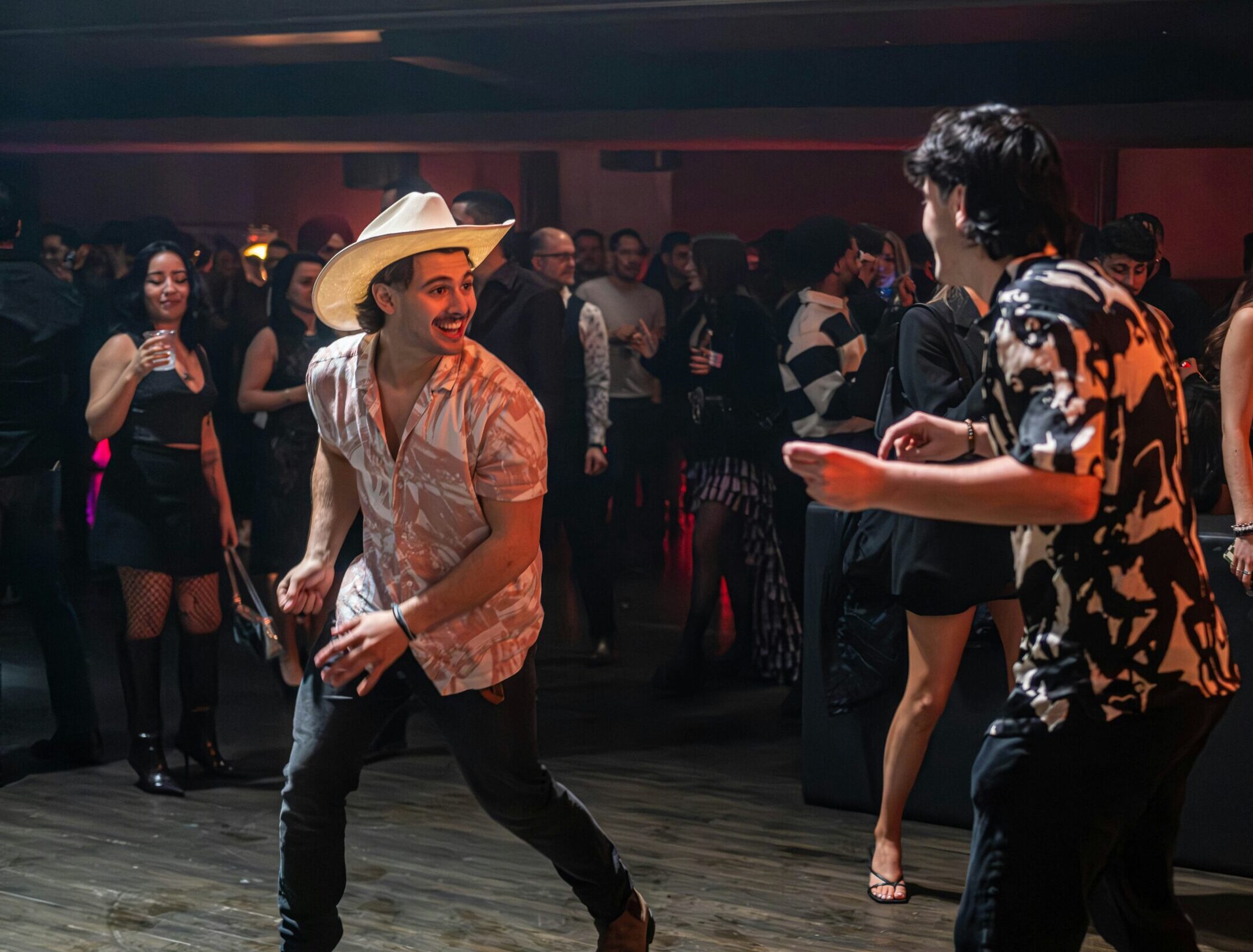 mehrere Tanzendende Menschen mit Cowboyhüten in einem dunklen Saal