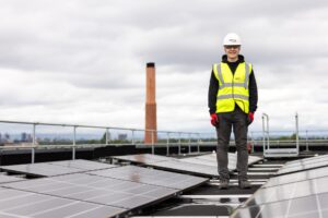 Junge steht in Arbeitsweste und Helm auf einem Dach zwischen Solarpanelen