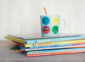 Auf dem Bild sieht man mehrre Kinderbücher übereinandergstapelt mit einer bunten Tasse die oben drauf steht