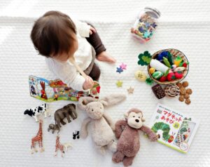 Auf deme Bild sieht man ein Kleinkind von oben fotografiert wie es vor eiener Auswahl an Spielsachen sitzt auf einer weißen Decke